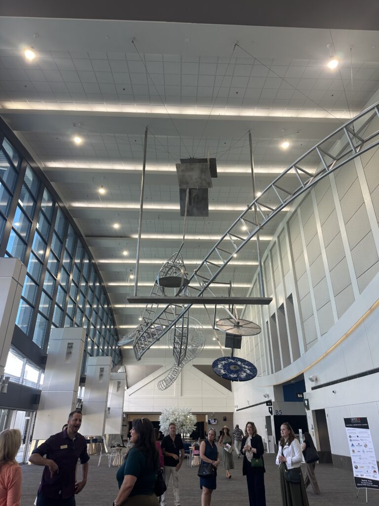 Modern convention center interior with a suspended abstract sculpture made of metal shapes and wires, and a group of attendees gathered below in conversation.
