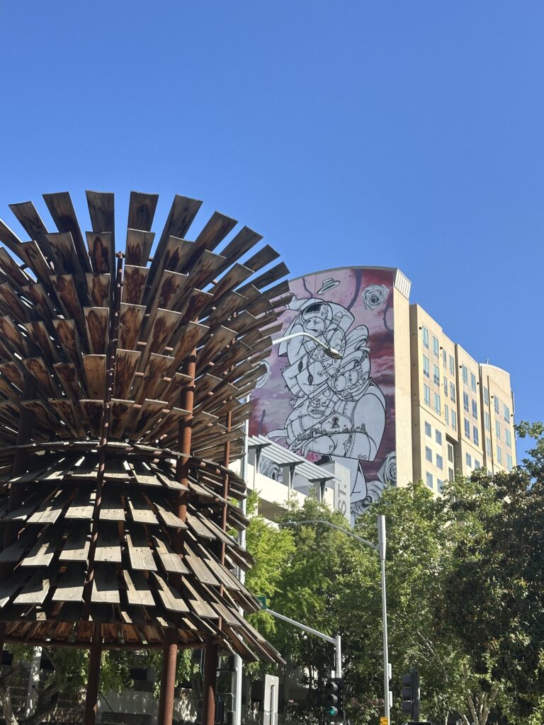 Outdoor public art installation featuring a wooden, cone-shaped sculpture in the foreground with a large building mural in the background depicting a stylized astronaut figure against a red and white backdrop.