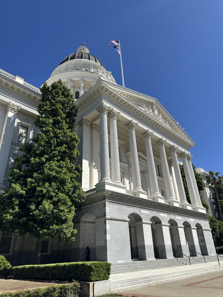 Exterior view of the California State Capitol building in Sacramento, with tall white columns, a central dome, and American and California flags waving in the blue sky. 