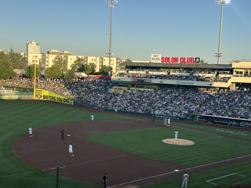 Crowded baseball stadium during a game at sunset, with players on the field and fans filling the stands. The scoreboard displays 'Solon Club' and other sponsor signs.
