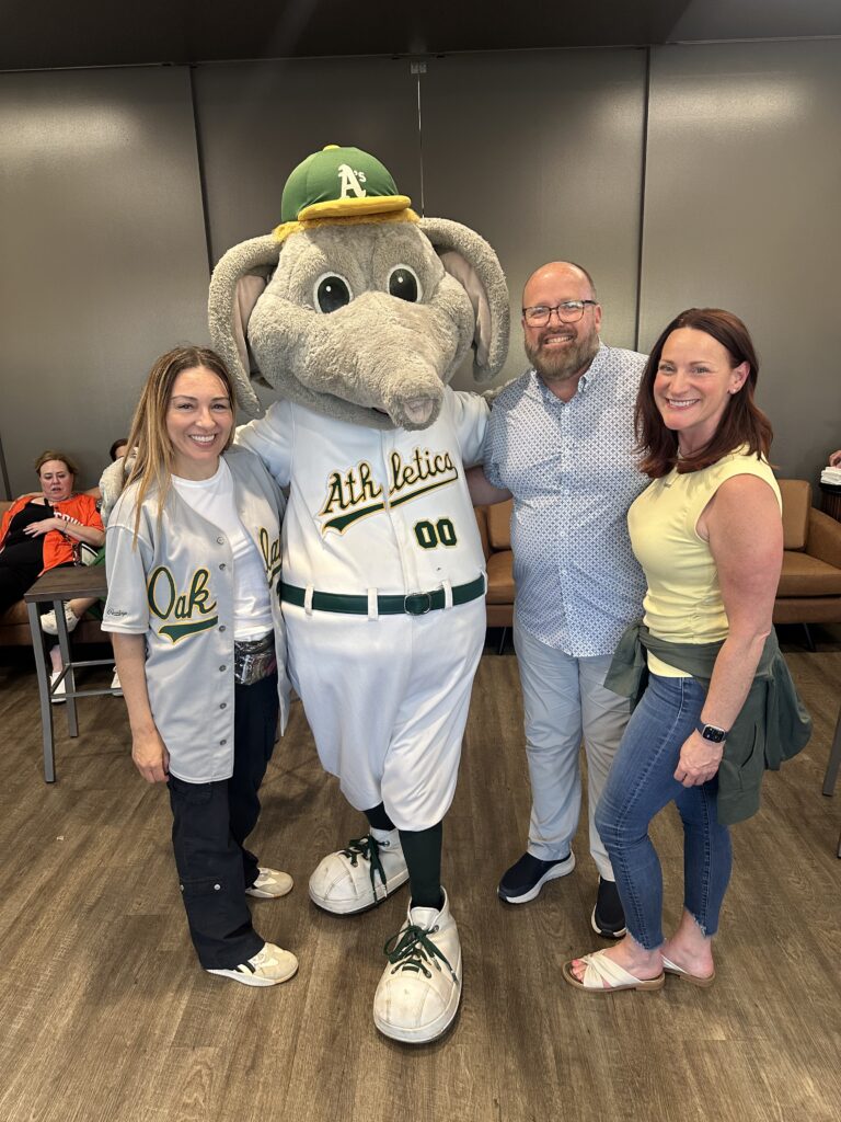 Three people posing and smiling with the Athletics' mascot, an elephant wearing a baseball uniform and green cap, inside a VIP suite area at a game.